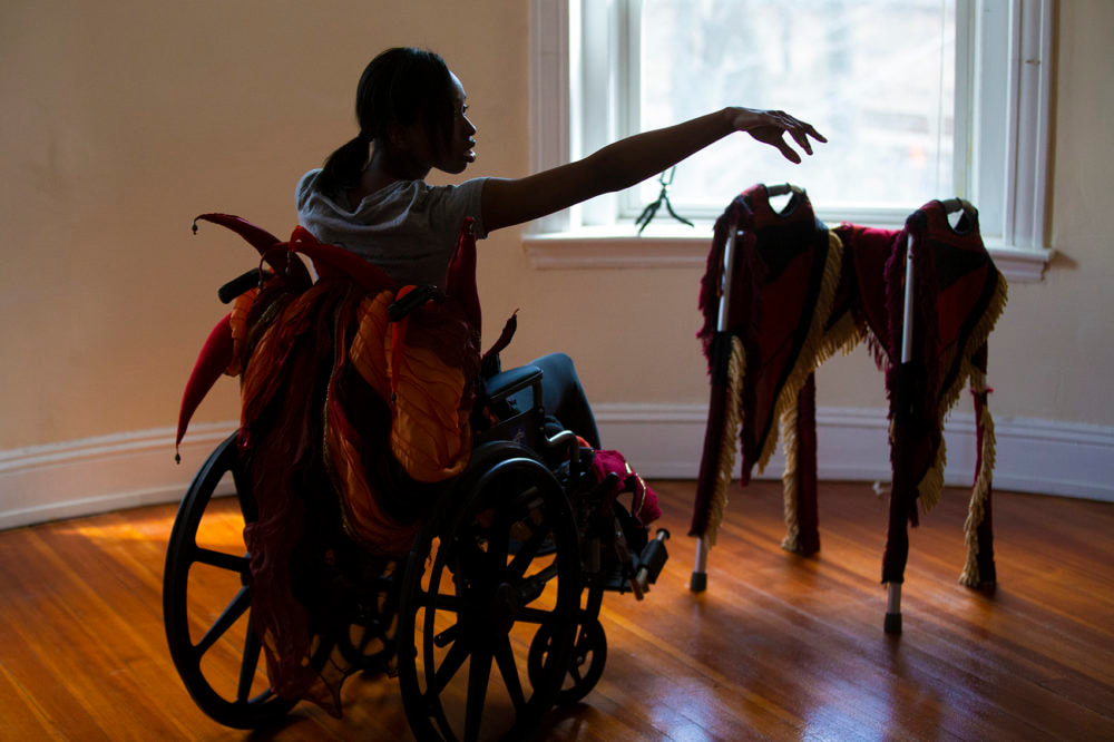On the left is a dark haired, dark skinned woman in a wheelchair with orange and red tendrils peeking up from the back of the chair. On the right, a heavily fringed walker sits in front of an open widow.