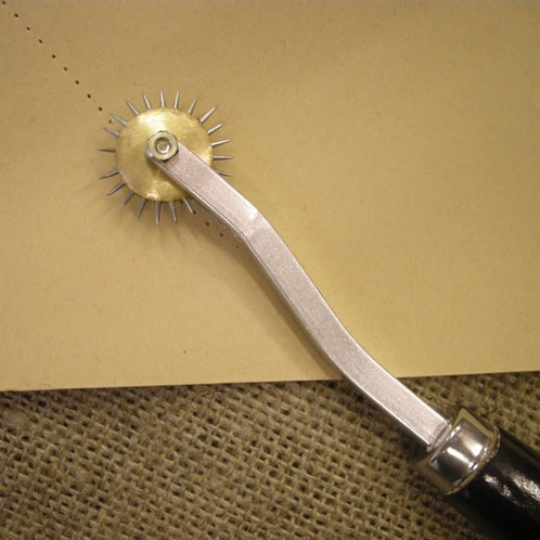 A spiky tracing wheel lays on paper. Its handle is wood and metal and the wheel on top has spikes all around it. There is a perforated line on the paper below the wheel.