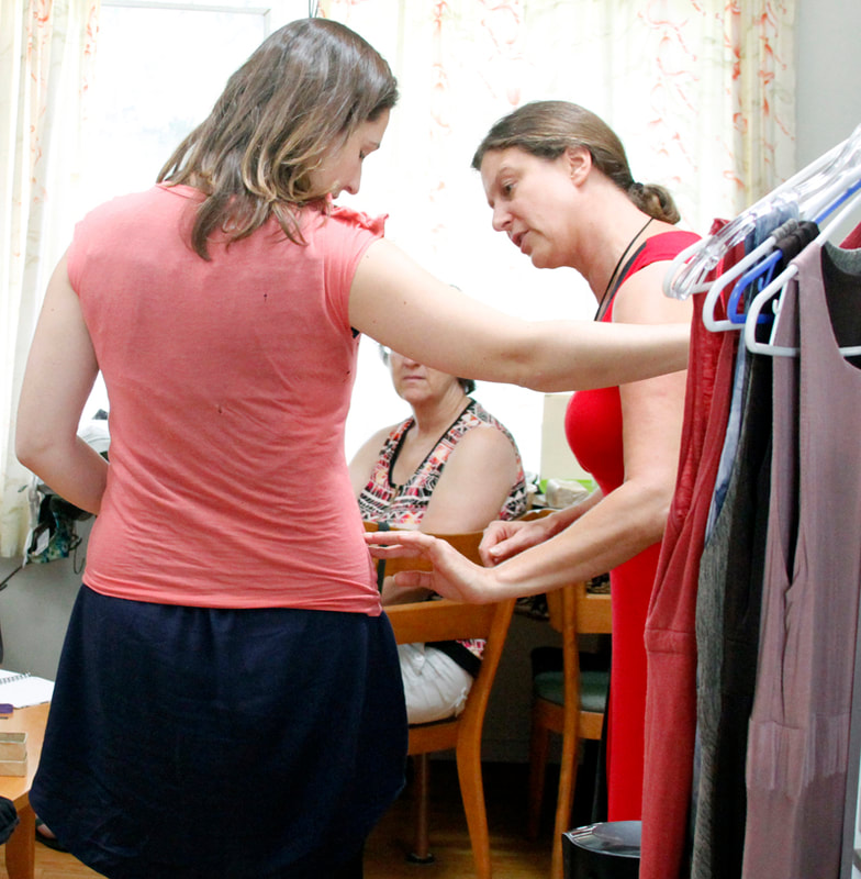 A woman with her back to us is being fitted into a pink t-shirt while a woman in red on the right points out the alterations.