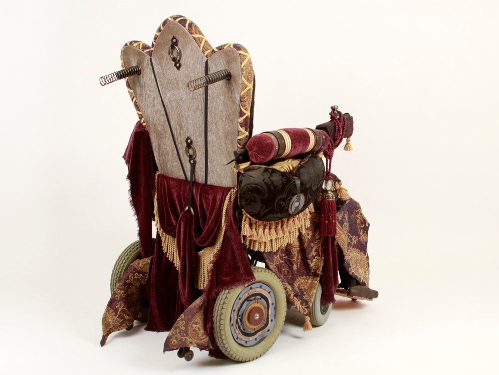 A wheelchair is reupholstered to look like an Edwardian throne. It has tapestry padding, purple drapery, golden fringe, and a silver metallic back with attached ball bearings.