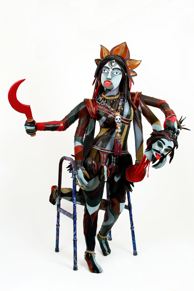 A sculpture representing the goddess Kali is built into a walker. The figure has 6 arms, 4 lower legs, and multi-colored skin. She holds a red scythe, a severed demon head, and a bowl.