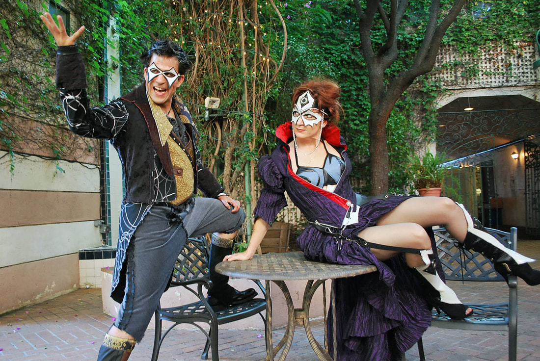 Two masked characters in colorful costumes tell stories around a cafe table in a wooded courtyard.