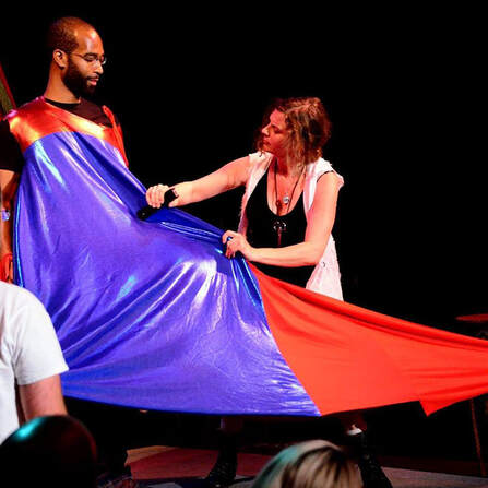 A dark skinned, bearded man with glasses wears a tent of shiny red and blue fabric. On the right, a light skinned woman in black and white staples the fabric around him.