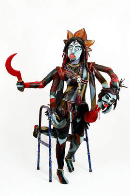 A version of the goddess Kali built into a walker. The sculpture has multicolored skin, six arms, four lower legs, and three eyes. She holds a red scythe in her left upper hand and a severed demon head in her right upper hand.