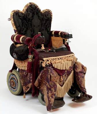 A wheelchair is reupholstered to look like an Edwardian throne. It has tapestry hangings, purple drapery, golden fringe, and a black tapestry padded seat edged with gold and purple.