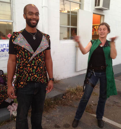 On the left, a bearded dark skinned man in glasses stands wearing a vest of chili print fabric. On the right is a woman in black with a long green vest smiling and waving her hands.