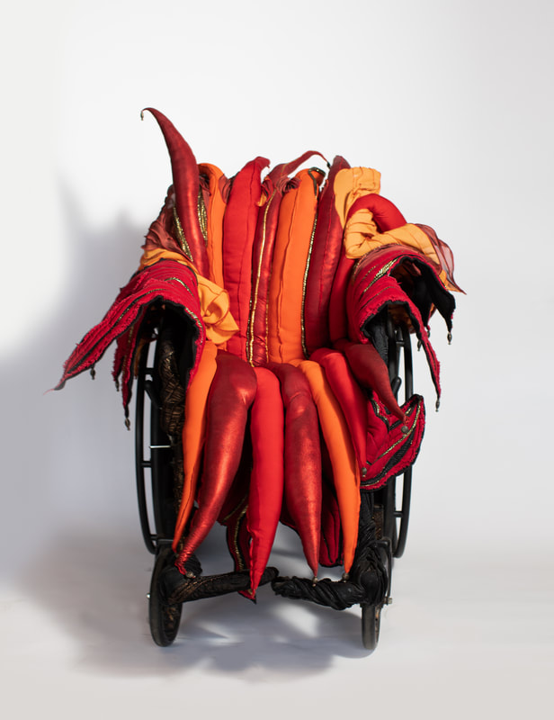 A wheelchair seat cover in red, orange, and gold padded spikes tipped with brass beads.