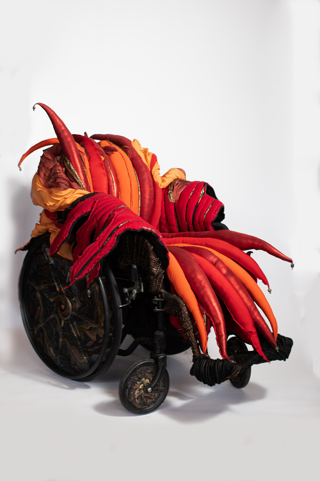 A wheelchair with copper, black, and multicolored swirls on its wheels has a seat cover covered in red, orange, and gold padded spikes tipped with brass beads.
