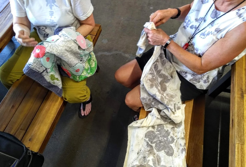 Two people sitting on benches, hand sewing patches onto fabric runners.