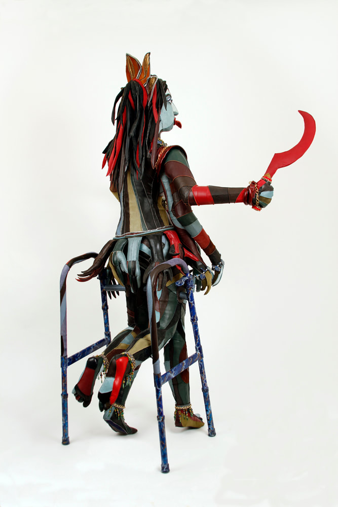 Back view of a sculpture representing the goddess Kali that is built into a walker. The figure has 6 arms, 4 lower legs, and multi-colored skin. She holds a red scythe in her top right hand.