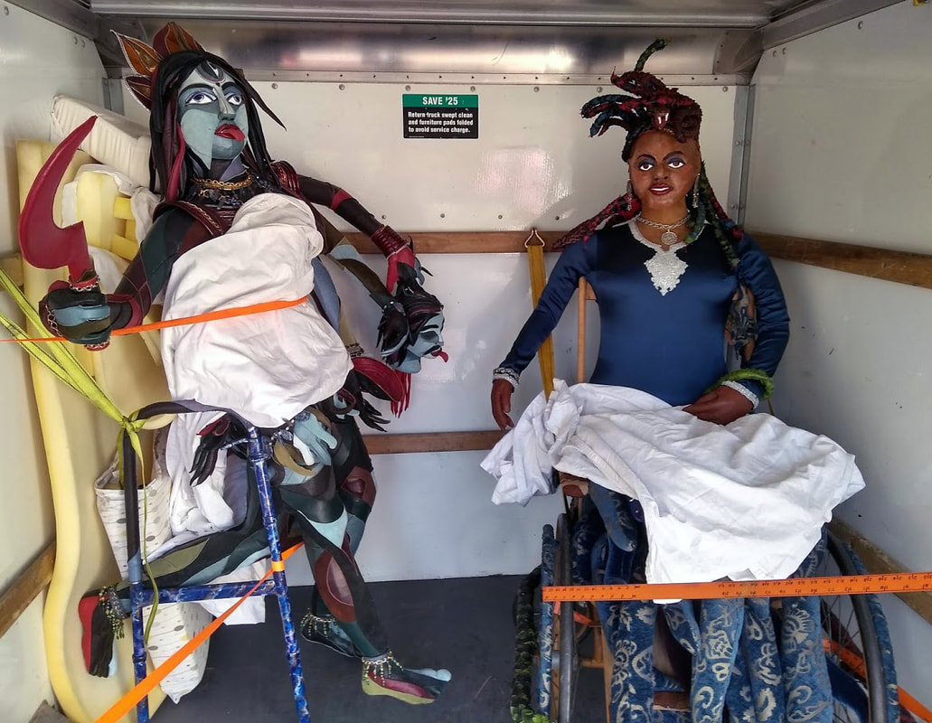 A sculpture of the goddess Kali built into a walker and a sculpture of Medusa built into a wheelchair are strapped into the back of a van and padded with sheets and foam.