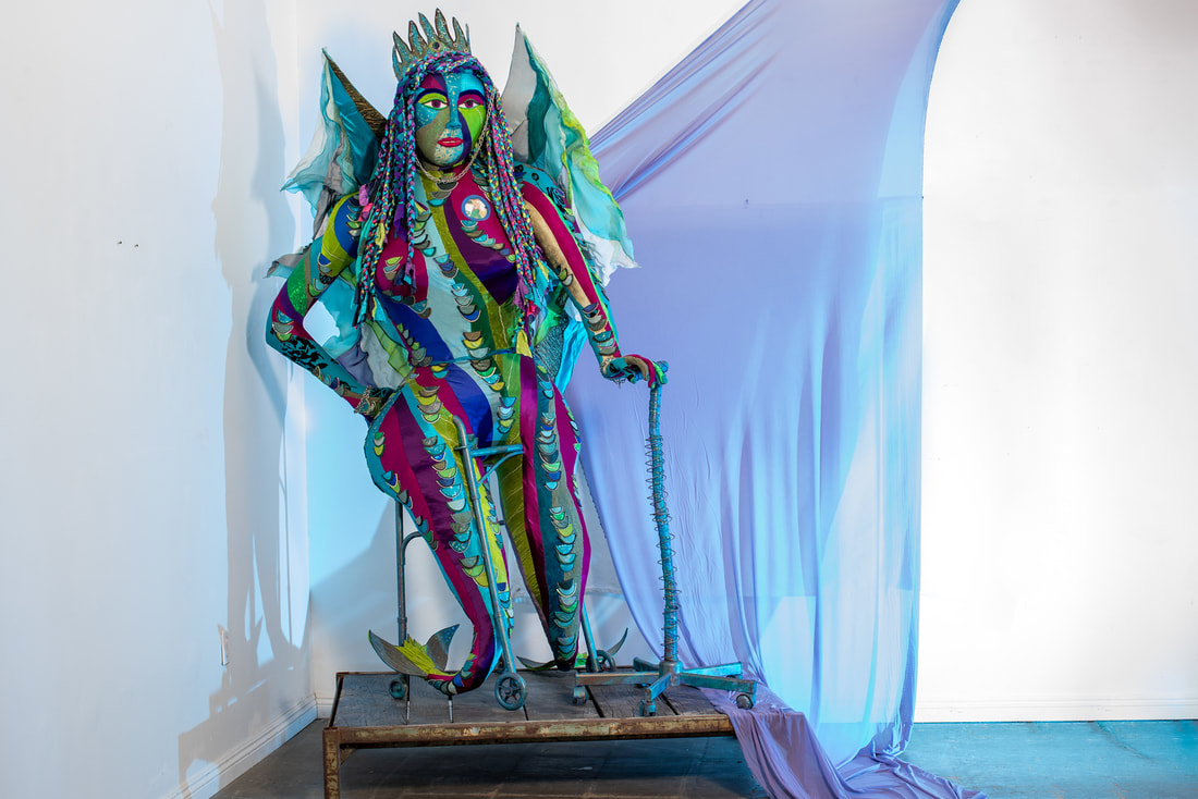 A sculpture of a winged multicolored woman with fishtail legs built into a walker. She holds a rolling cane with her left hand and rests her right fist on her right hip.