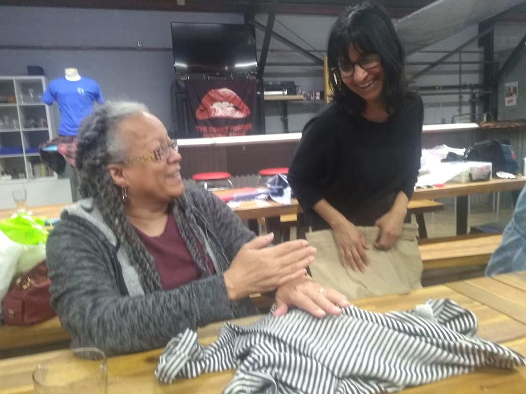 Two women in glasses are  working at a table. They are smiling and holding clothing to repair.