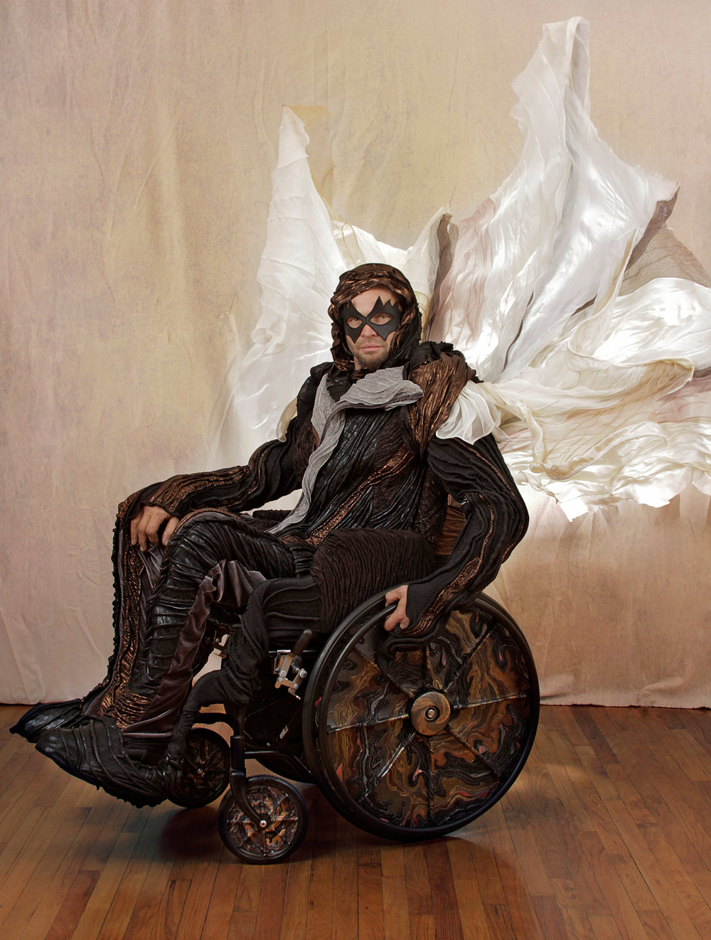 Left side view of a masked man in black, copper, and silver drives a wheelchair with a white and silver sail. The chair's wheels are covered in black, copper, and multi-colored metallic swirls.