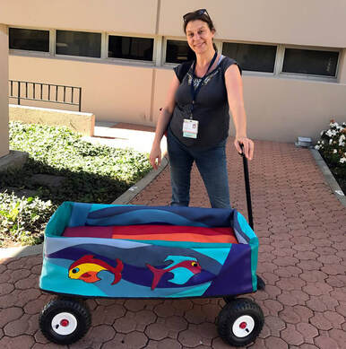 A light skinned woman in jeans and a t-shirt stands, holding the handle of a wagon dressed in a vinyl cover with brightly colored fish and waves.