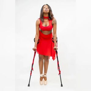 A brown skinned dander in a red top and skirt holds hersolf up on her toe shoes with red wrapped crutches