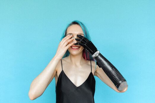 Against a sky blue background is a pae skinned woman with blue hair and a black slip. She has one black prosthetic arm and hand, and holds her hand and the prosthetic hand up over her face, covering her eyes. 