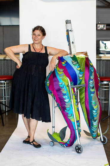 A smiling pale skinned woman in black leans against a sculpture of a pair of colorful legs/fish tails built into a walker.