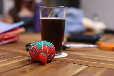 A tomato-shaped pincushion sits in front of a pint glass of dark lager on a picnic table.