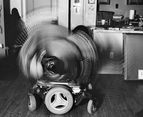 A black and white photo of a wheelchair base on a hardwood floor. Above the base, the chair tilts back and forth creating a blurred semi-circle. In the backgroun is a dark shadow of a figure on the left and kitchen counters on the right.