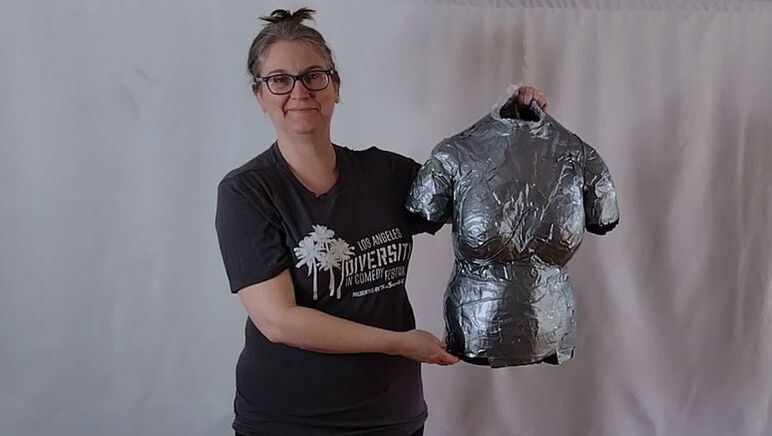 A smiling woman with glasses wearing a grey T- shirt holds up a silver duct tape dress form.