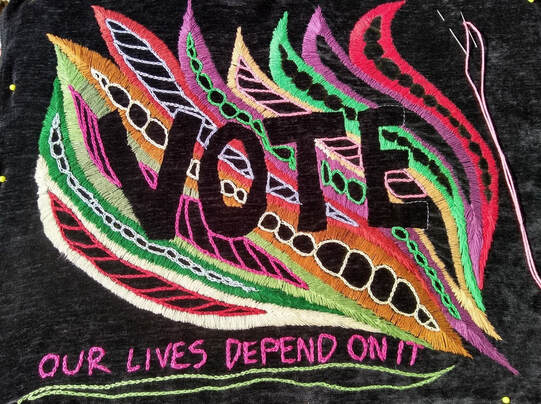 Colorful embroidery outlines the word VOTE in black. The words OUR LIVES DEPEND ON IT are embroidered in pin at the bottom.