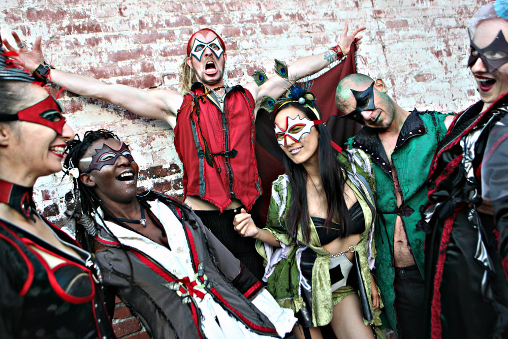 6 masked people in colorful costumes laugh and gesture in front of a brick wall.