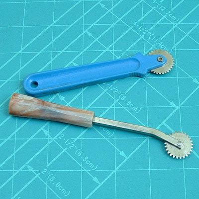 Two tracing wheels lay on a turquoise cutting mat. They have plastic handles and dull serrated wheels at the top.