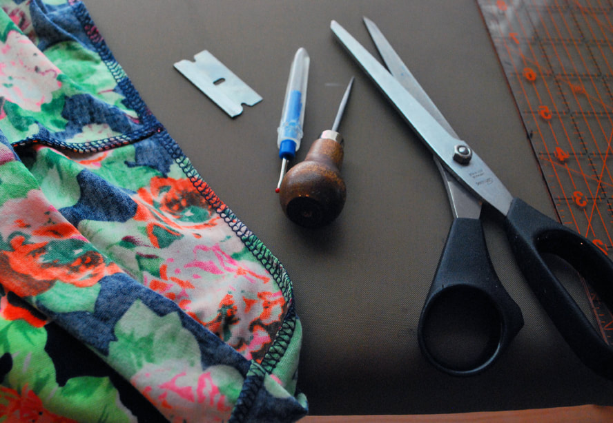 A dress printed with bright colored flowers next to a safety blade, a seam ripper, an awl, and a pair of scissors.