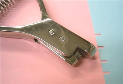 A curved metal tool lays on a pink surface next to blue paper. The paper has 4 even notches cut out of one side.