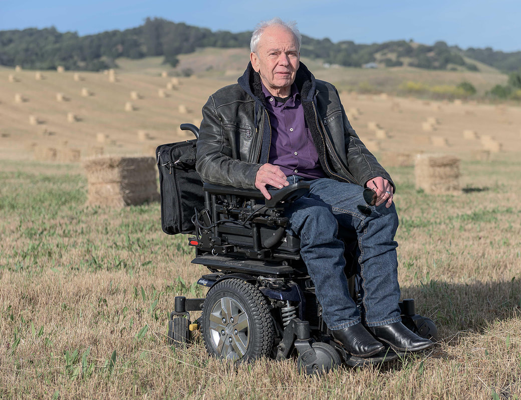 Balding pale skinned man in jeans, a purple shirt, and a black leather jacket sits in his wheelchair in a field full of hay bales.