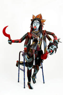 A version of the goddess Kali built into a walker. The sculpture has multicolored skin, six arms, four lower legs, and three eyes. She holds a red cured sword in her left uipper arm and a severed demon head in her top right hand.