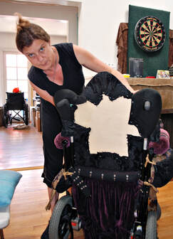 A woman with a needle in her mouth bends over the back of a wheelchair. She is reupholstering the chair's back with tapestry fabric.