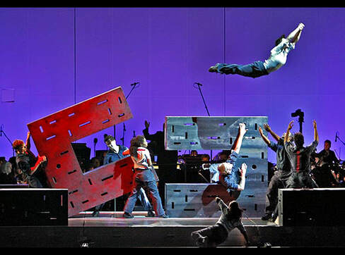 A dancer hurls herself over huge red and blue blocks toward the waiting arms of her fellow dancers.