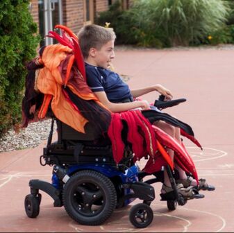 A boy with blonde hair drives an electric wheelchair. The seat of the chair is covered in red, orange, gold, and peach tendrils with beaded tips.