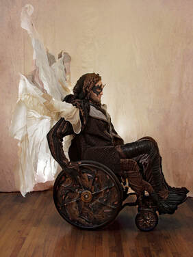 Right side view of a masked man in black, copper, and silver drives a wheelchair with a white and silver sail. The chair's wheels are covered in black, copper, and multi-colored metallic swirls.