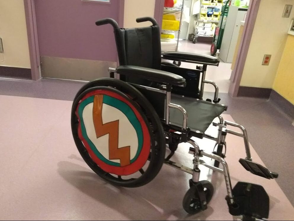 A classic manual wheelchair has a vinyl wheel cover in red, white, black, and green with a metallic orange lightening bolt down the center.