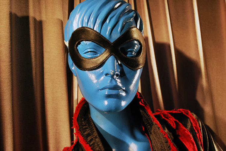 A blue store mannequin face and shoulders wearing a black mask and a coat with a red and black collar.