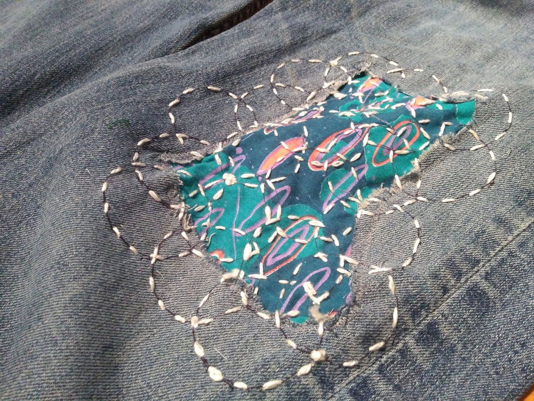 Visible mending on a pair of faded blue jeans: ciruclar stitching in white over a colorful blue and orange patch.