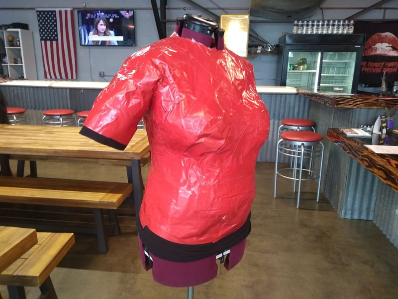 A mannequin made of bright red duct tape stands in front of picnic tables and bar stools.