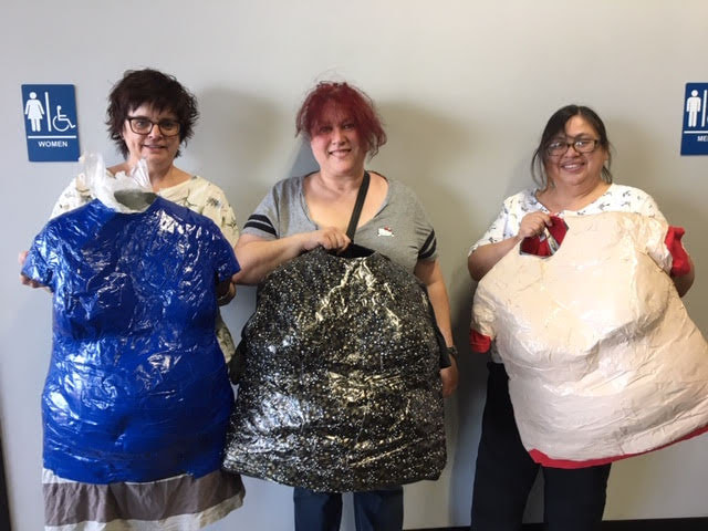 Three smiling women hold up duct tape dress forms: one in blue, one black with silver stars, and one in cream.