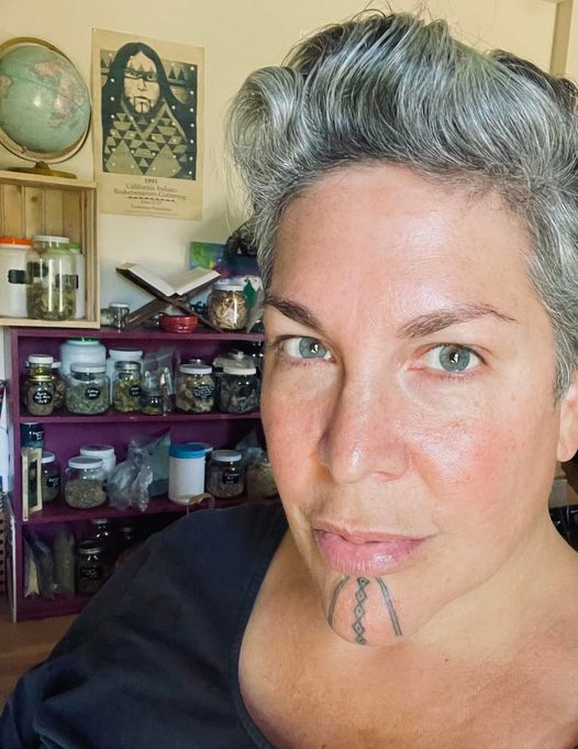 A woman with short grey hair, grey-blue eyes, and blue chin tattoos in front of an apothecary cabinet.