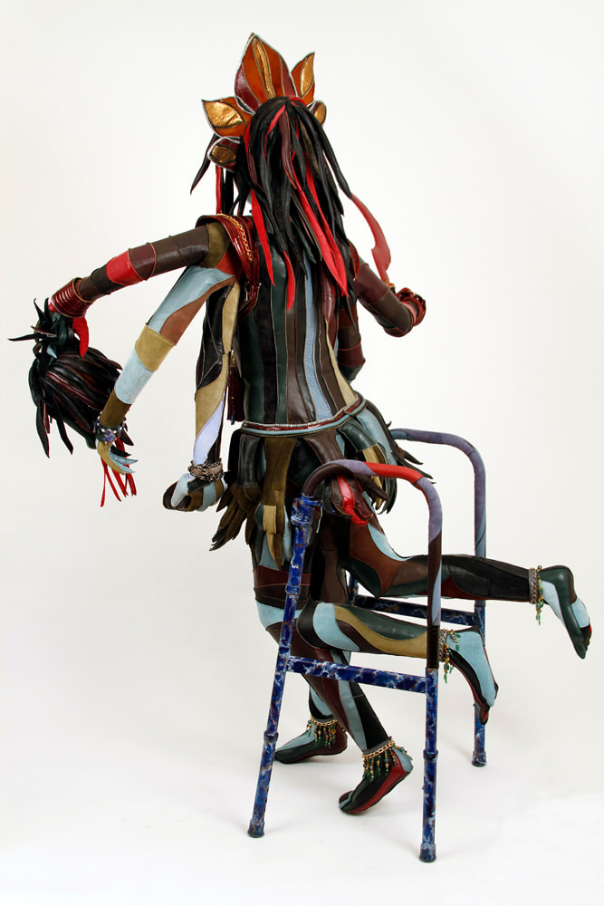 Back view of a sculpture representing the goddess Kali that is built into a walker. The figure has 6 arms, 4 lower legs, and multi-colored skin. She holds a severed demon head in her middle left hand.