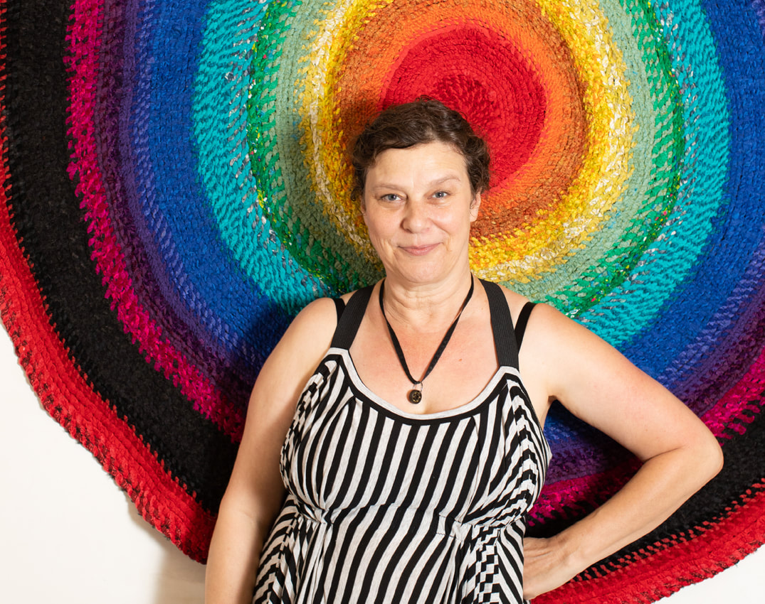 A smiling woman in a grey and black striped dress in front of a rainbow colored rag rug.