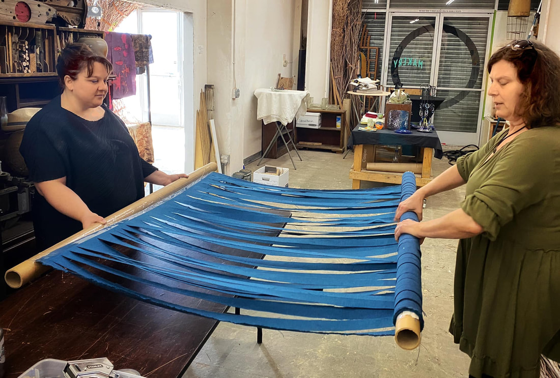 Two brown haired women stand apart, holding cardboard tubes with a row of strips of bright blue fabric taped to both tubes. In the background is an artist studio.
