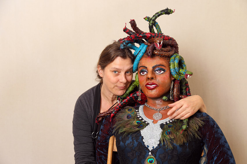 A white woman with light brownhair puts her arm around a sculpture of Medusa. Medusa has snakes for hair and wears a peacock feather necklace.