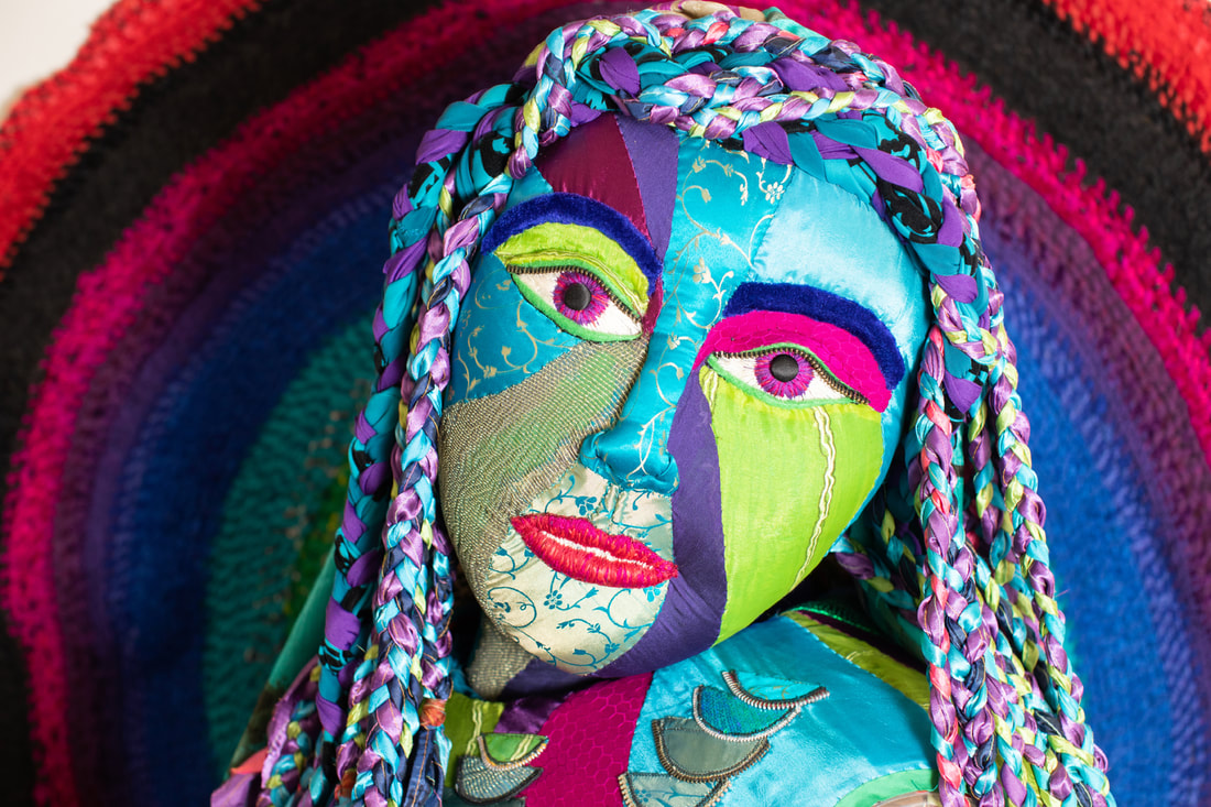 A scupted female face covered in green, blue, violet, gold, and purple fabric with embroidered lips and eyes.