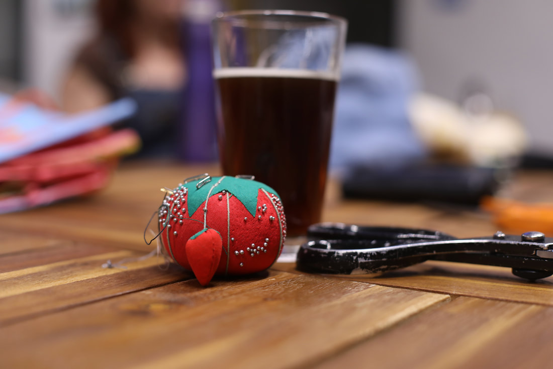 A pincushion and scissors sit on a table in front of a nearly full glass of porter.