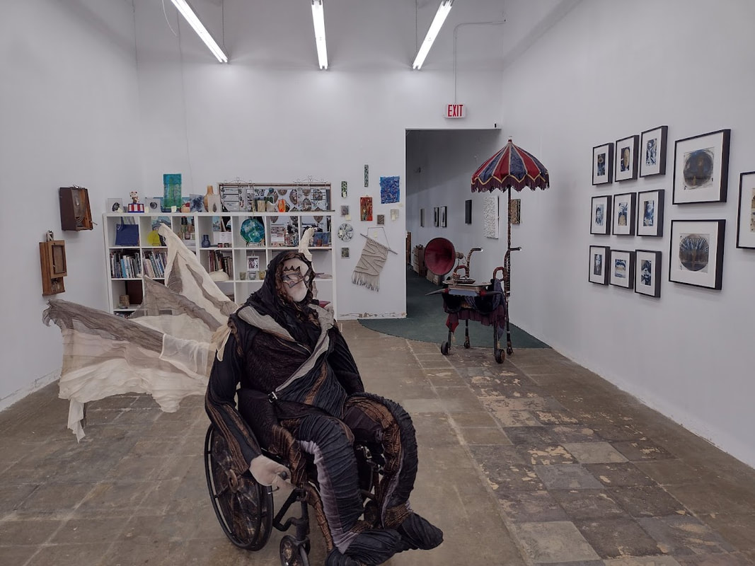 Sculpture of a masked figure in black and copper in a matching wheelchair and a Victorian style walker with attached parasol, gramophone horn, and cowhorns in front of white walls hung with art.