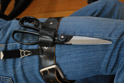 A leg in medium blue denim wearing a black leather thigh holster which holds a large pair of scissors.